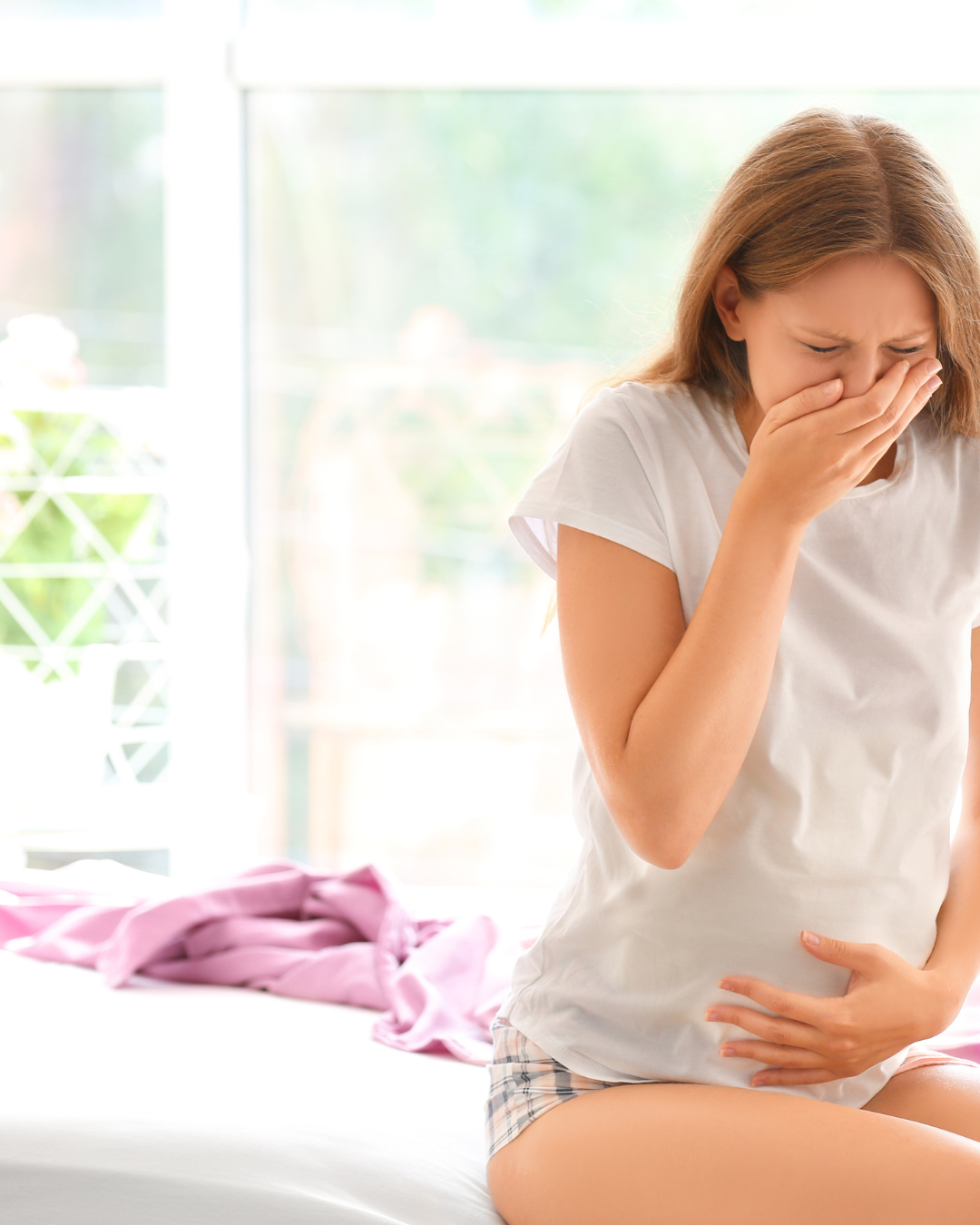 Pregnant woman covering her mouth and holding her stomach, appearing nauseous in a bright bedroom.